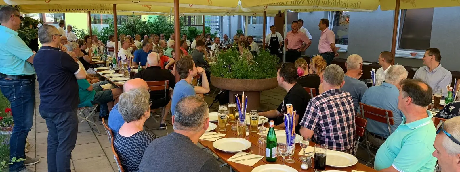 Das Bild zeigt einen vollbesetzten Biergarten. Im Hintergrund stehen Menschen die eine Ansprache halten.